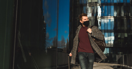 Business trip during coronavirus pandemic. Young man with luggage case against the background of the airport or train station building. Wearing mask and social distancing on city street public place