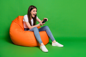 Photo of excited funny brunette hair young woman play playstation wear t-shirt jeans sneakers sit on pouf isolated on green background