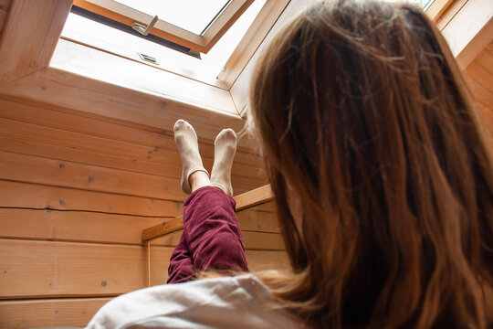 A young girl sits on Chrisla putting her foot on her leg against the background of an attic window in the attic of a wooden background