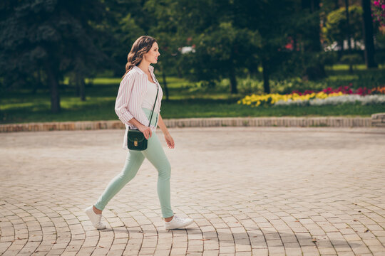 Full size profile photo of cute brunette lady walk park active life wear pastel green jeans white top striped shirt shoulder bag outdoors