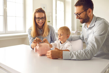 Little boy with his parents puts coins in a piggy bank sitting at a table in the room.