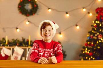 Happy boy in traditional Christmas jumper and Santa cap sitting at table and smiling