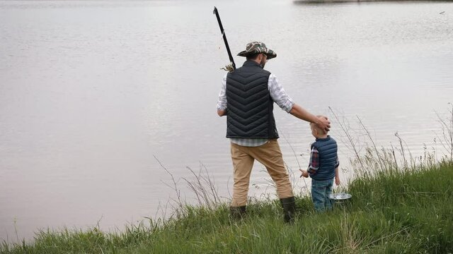Man with a fishing rod on his shoulder and little son are standing on the shore of the lake. Dad and son go fishing