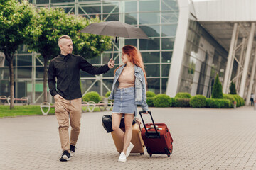 Young couple, man and woman near the airport with a suitcase. Happy couple traveling. Young people returning from a trip on a rainy day, a man holds an umbrella