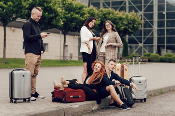 Five friends waiting for the flight, sitting on the asphalt, using mobile phones. Three girls and two boys with suitcases go on a trip