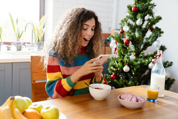 Excited charming woman using mobile phone while having breakfast