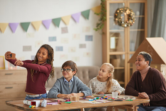 Waist up portrait of teenage African-American girl taking selfie with kids and teacher during art and craft class, copy space