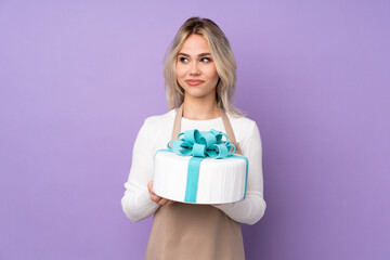Young pastry chef holding a big cake over isolated purple background standing and looking to the side