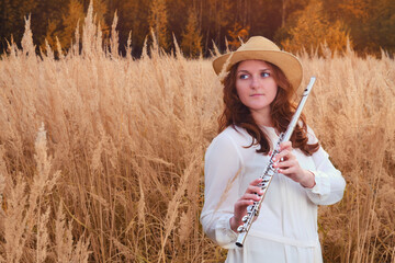 A young woman in straw hat with red hair plays a flute in an autumn field. A musician with a wind...
