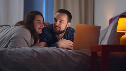 Cheerful young couple looking at laptop screen web surfing together in bedroom