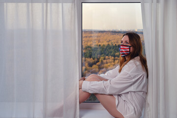 Woman in medical mask with usa flag sits on windowsill by window. Concept of coronavirus quarantine and home isolation