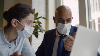 Business people with face masks working together on laptop in modern coworking office