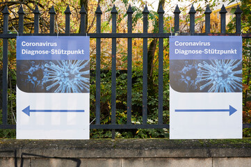 Wegweiser zum Coronavirus-Diagnose-St&uuml;tzpunkt am Zaun in beide Richtungen