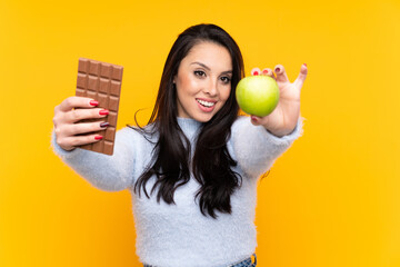 Young Colombian girl over isolated yellow background taking a chocolate tablet in one hand and an...