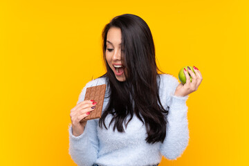 Young Colombian girl over isolated yellow background taking a chocolate tablet in one hand and an...