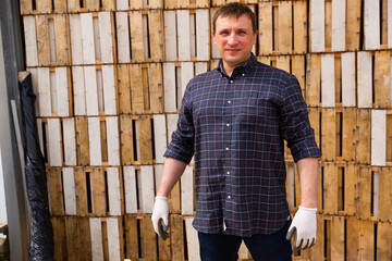 Portrait of confident adult farmer standing on background of stack of slatted wooden cases for future harvest