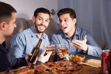Three positive men talking and laughing while enjoying beer and pizza at home