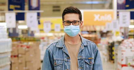 Close up portrait of happy young Caucasian male in mask and glasses standing at supermarket...