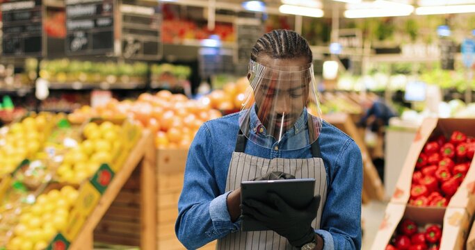 Close up portrait of happy African American male manager in face shield standing at supermarket and typing on tablet. Joyful salesman in apron working and browsing on device in grocery. Retail concept