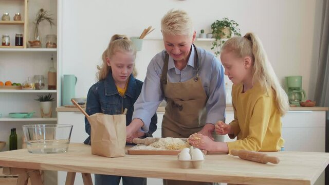 Medium of good-looking Caucasian granny teaching cooking two twin blond granddaughters, making and kneading dough. Family cooking, talking, having fun in kitchen together