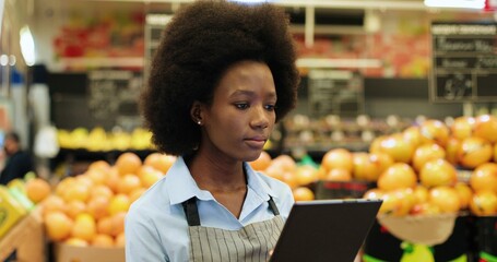 Portrait of joyful female grocery store assistant in apron holding packet with fresh organic vegetables, smiling to camera in good mood. Young African American woman at supermarket. Retail concept