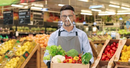 Portrait of joyful Caucasian male food store manager in face shield and gloves standing in supermarket and holding box with fresh vegetables, smiling at camera. Handsome guy seller working at grocery.