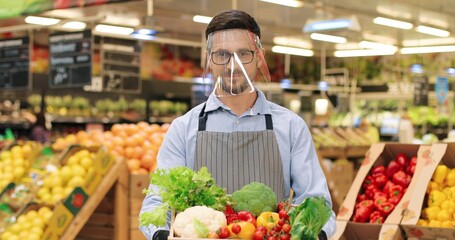 Portrait of joyful Caucasian male food store manager in face shield and gloves standing in supermarket and holding box with fresh vegetables, smiling at camera. Handsome guy seller working at grocery.