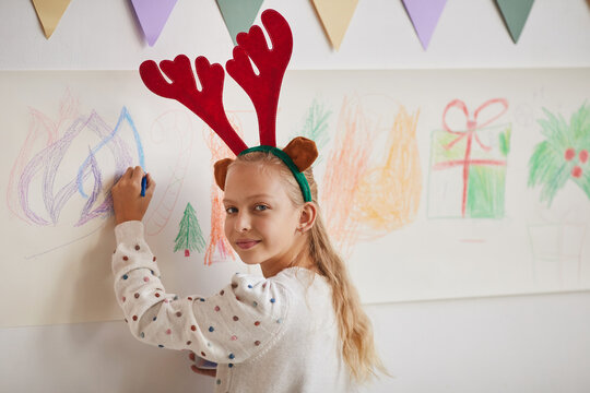 Portrait of smiling blonde girl wearing antlers drawing Christmas pictures on wall during art class, copy space