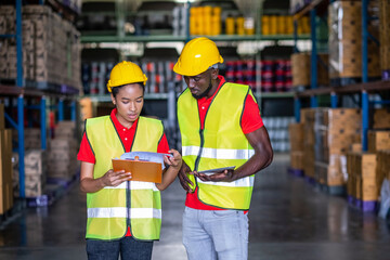 Africa Americans using a tablet and standing with an engineer woman hold clipboard and check stock in the automotive parts warehouse. Wear a safety helmet and vest. In background shelves with goods
