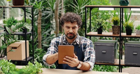 Handsome young male florist in apron tapping on tablet sitting at table in own flower shop. Happy Caucasian man manager browsing on device in floral greenhouse. Small business concept