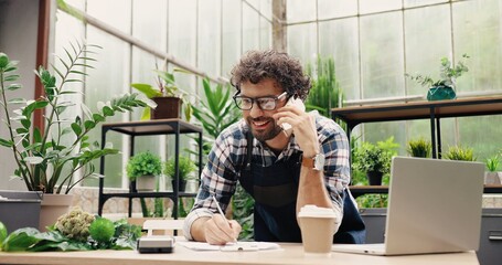 Happy Caucasian businessman talking on cellphone while standing in apron in small floral center and writing down order details. Joyful male florist calling on smartphone at work. Own business concept