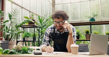 Happy Caucasian businessman talking on cellphone while standing in apron in small floral center and writing down order details. Joyful male florist calling on smartphone at work. Own business concept - obrazy, fototapety, plakaty