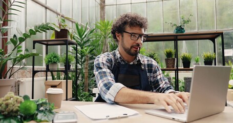 Portrait of Caucasian man entrepreneur in glasses sitting in own flower shop, typing on laptop and...