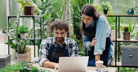 Portrait of happy Caucasian male and female florists flower store owners working and doing inventory. Man typing on laptop at desk in floral shop and talking to woman boss. Business concept