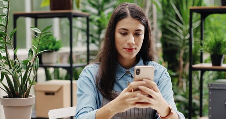 Close up portrait of cheerful Caucasian woman in good mood texting on smartphone in own floral center. Pretty happy female florist at work in flower store typing on mobile phone. Business concept