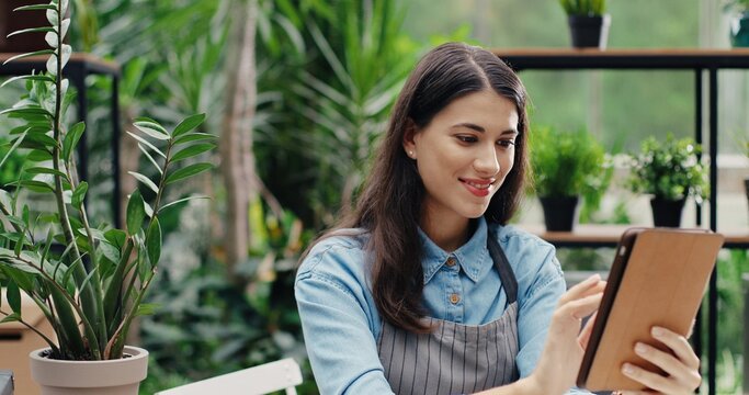 Close up of young Caucasian beautiful female florist entrepreneur in apron sitting at desk and typing on tablet. Portrait of woman worker in flower shop tapping on device. Floral business concept
