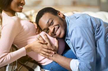 Happy black man hugging his pregnant girlfriend's big belly and listening to unborn baby's heartbeat at home
