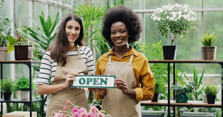 Happy mixed-races women florists in aprons standing in own floral shop and smiling to camera with Open sign in hands. Caucasian and African American females reopening flower store. Portrait concept