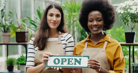 Close up portrait of joyful mixed-races female employees in aprons standing in floral shop holding Open sign in hands and smiling to camera. Caucasian and African American females opening flower store