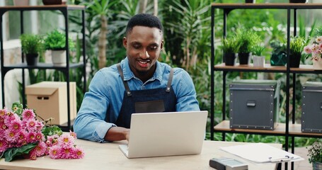 Joyful young man in flower shop sitting at workplace and texting on computer. Cheerful African American male florist worker in floral house tapping and typing on laptop. Floristry concept
