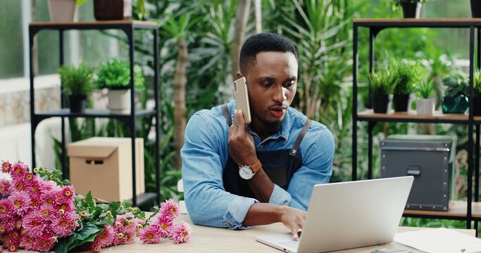 Young African American man entrepreneur sitting at desk and typing on computer while chatting on smartphone. Happy male florist in green floral center tapping on laptop and calling on cellphone