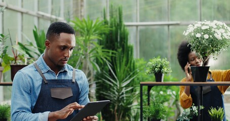 Close up portrait of joyful African American man florist typing on tablet while doing flower orders at flower shop. Busy female watering flowers on background and calling on smartphone. Retail concept