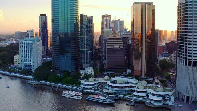 Golden Sunset Beaming By High-rise Buildings With Eagle Street Pier Ferry Terminal In Foreground At Brisbane River In Queensland, Australia. - aerial drone shot