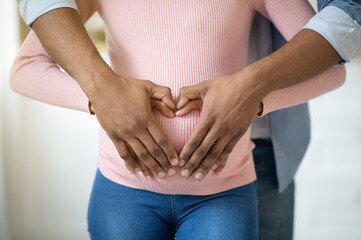 Closeup view of black pregnant woman and her husband hugging tummy together, making heart with their hands