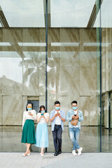 Group of young people in protective masks standing at building wall outdoors and texting friends or checking social media