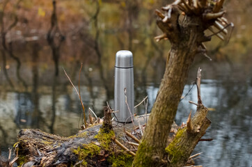 Vacuum steel thermos outdoors. Aluminum hiking flask, forest and pond in the background. Hike concept, relaxation in nature, hot drink in cold weather.