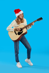 Young woman in Santa hat playing acoustic guitar on light blue background. Christmas music