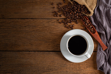 Top view above of Black hot fresh coffee in a white ceramic cup with coffee beans roasted in burlap sack bag on wooden table background. Flat lay with copy space.