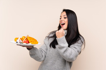 Young Colombian girl holding waffles over isolated background celebrating a victory