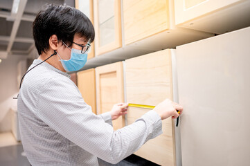 Asian man wearing face mask using tape measure on cabinet panel choosing wooden materials for...